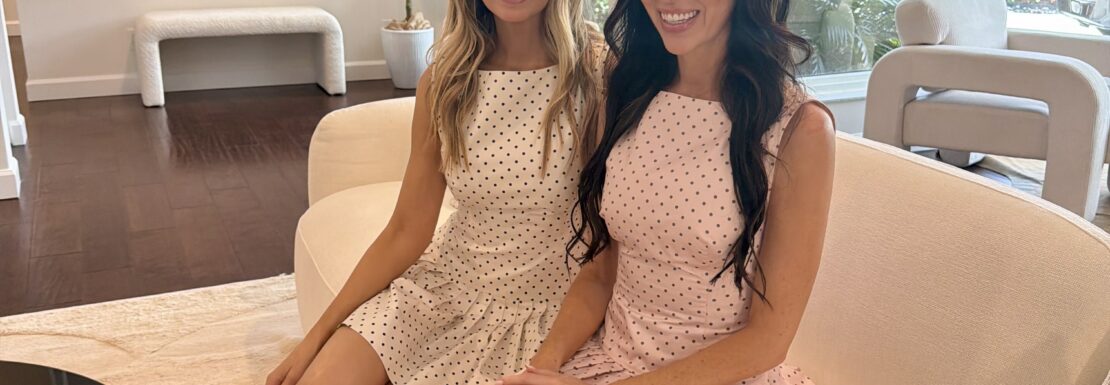 Professional portrait of realtors Ashleay Vesper and Heather Costa of The Heather Ashleay Team sitting on a white sofa in a modern, brightly lit Fort Lauderdale home.