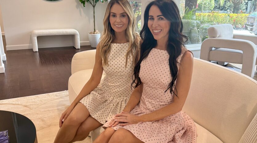 Professional portrait of realtors Ashleay Vesper and Heather Costa of The Heather Ashleay Team sitting on a white sofa in a modern, brightly lit Fort Lauderdale home.